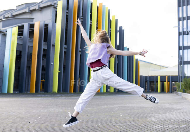Полная длина счастливая молодая пригодная женщина в повседневной одежде прыгая с поднятыми руками против творческого современного здания и глядя прочь с улыбкой на ясную летнюю погоду — стоковое фото