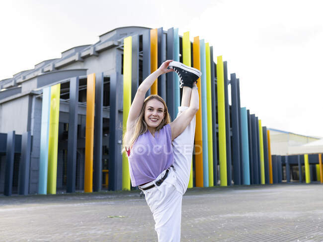 Full length attractive fit female in casual wear performing standing split and looking at camera with smile while standing on against creative building on sunny summer day — Stock Photo