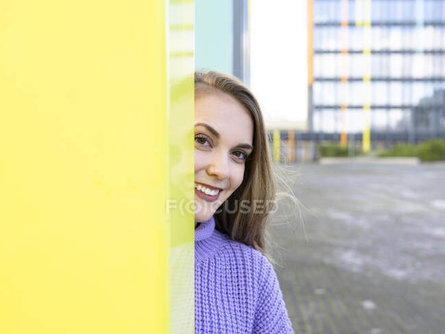 Привлекательная молодая женщина в голубом свитере стоит за желтой стеной на улице и смотрит в камеру с очаровательной улыбкой — стоковое фото