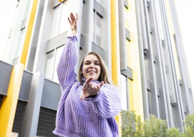 Sonriente joven hembra en suéter violeta levantando los brazos con gracia mientras está de pie contra el edificio contemporáneo y mirando a la cámara felizmente - foto de stock
