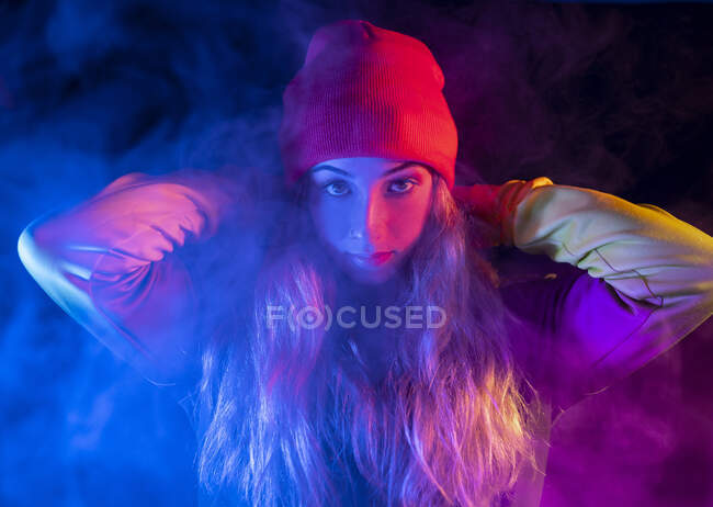 Молодая уверенная в себе леди с длинными светлыми волосами в стильном наряде и шляпе, стоящая в темно-дымчатой студии с неоновой подсветкой и смотрящая в камеру — стоковое фото