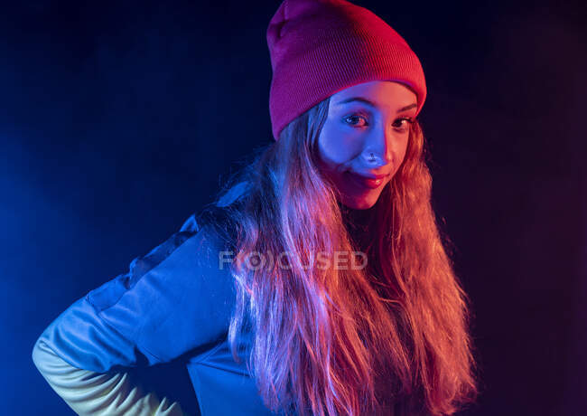 Young confident lady with long blond hair in stylish outfit and hat standing in dark smoky studio with neon illumination and looking at camera — Stock Photo