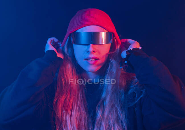 Модні молоді жінки тисячоліття з довгим світлим волоссям у футуристичних сонцезахисних окулярах регулюють капелюх, стоячи в темній кімнаті з неоновим освітленням — стокове фото