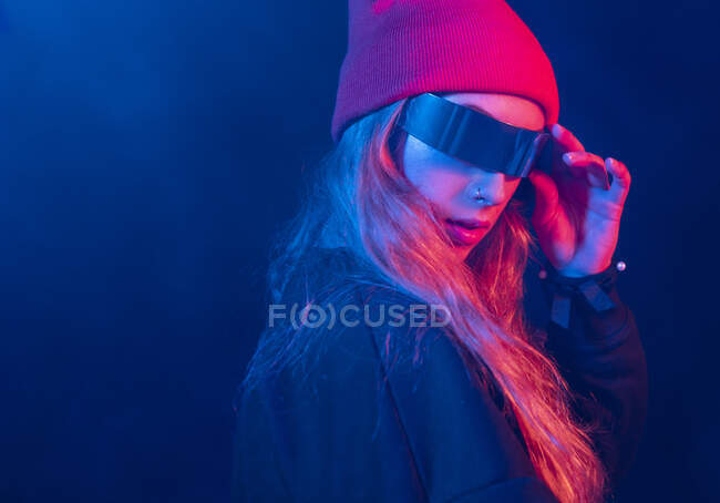 Trendy young female millennial with long blond hair in futuristic sunglasses adjusting hat while standing in dark room with neon illumination — Stock Photo