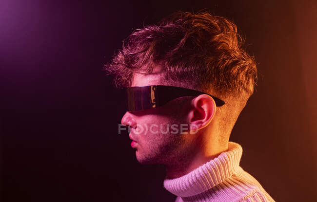 Side view of self assured young male in stylish outfit adjusting futuristic sunglasses while standing in dark studio with neon illumination — Stock Photo