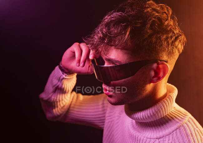 Боковой вид самоуверенного молодого мужчины в стильном наряде, регулирующем футуристические солнцезащитные очки, стоя в темной студии с неоновой подсветкой — стоковое фото