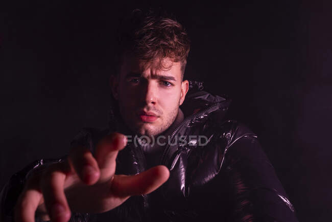 Thoughtful young unshaven guy in trendy outfit reaching out hand towards camera against black background — Stock Photo