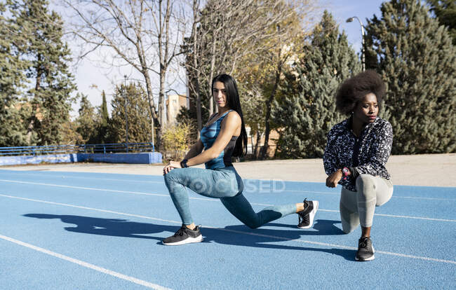 Fit multiracial female professional athletes in sportswear doing lunge exercise while warming up muscles during workout at stadium — Stock Photo