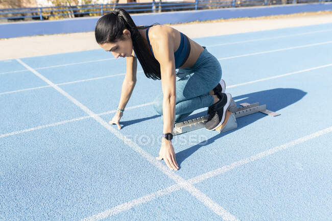 Vista lateral de la deportista de pie en los bloques de partida en la posición de inicio agacharse durante el entrenamiento en el estadio en verano - foto de stock