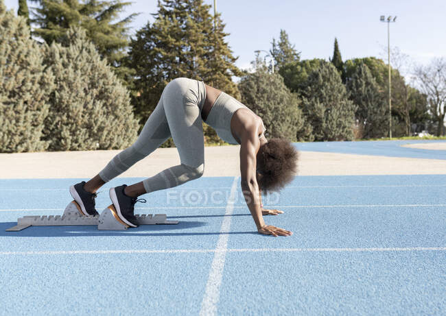 Вид сбоку афроамериканской бегуньи в стартовых блоках, стоящей в положении приседания, готовясь к спринту на стадионе во время тренировки — стоковое фото