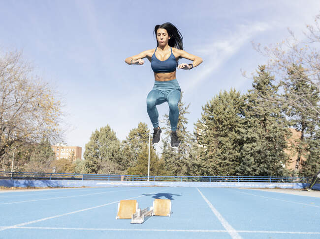 Determined female runner jumping above track with starting blocks while preparing for race at stadium during training — Stock Photo