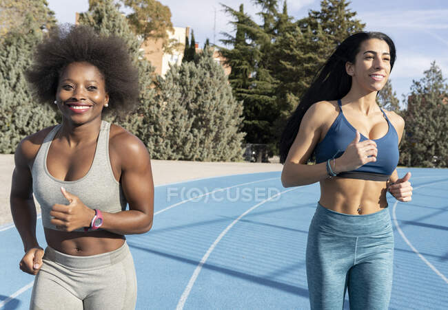 Corredores femininos multirraciais em sportswear correndo juntos no estádio enquanto sorrindo e desfrutando de treino no dia ensolarado — Fotografia de Stock