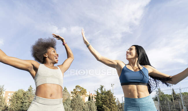 From below of delighted multiethnic female athletes in sportswear in moment of jumping above stadium track giving high five and looking at each other — Stock Photo