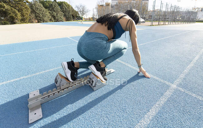 Side view of sportswoman sitting in starting blocks in crouch start position during training at stadium in summer — Stock Photo