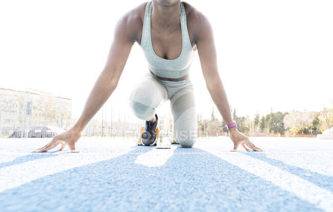 De baixo de afro-americano feminino corredor em blocos iniciais sentado na posição de agachamento, enquanto se prepara para sprint no estádio durante o treino — Fotografia de Stock