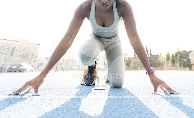 De baixo de afro-americano feminino corredor em blocos iniciais sentado na posição de agachamento, enquanto se prepara para sprint no estádio durante o treino — Fotografia de Stock