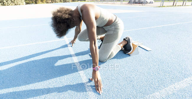 Сверху афро-американская бегунья в стартовых блоках, стоящая на корточках, готовясь к спринту на стадионе во время тренировки — стоковое фото