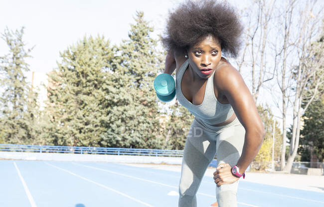 Focused black female athlete doing exercises with dumbbell while training at stadium in summer and looking forward — Stock Photo