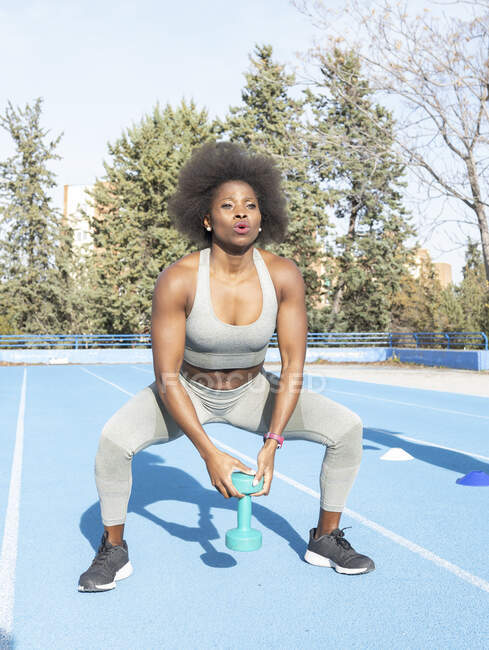 Atleta femenina negra enfocada haciendo ejercicios con mancuerna mientras entrena en el estadio en verano y mirando hacia adelante - foto de stock
