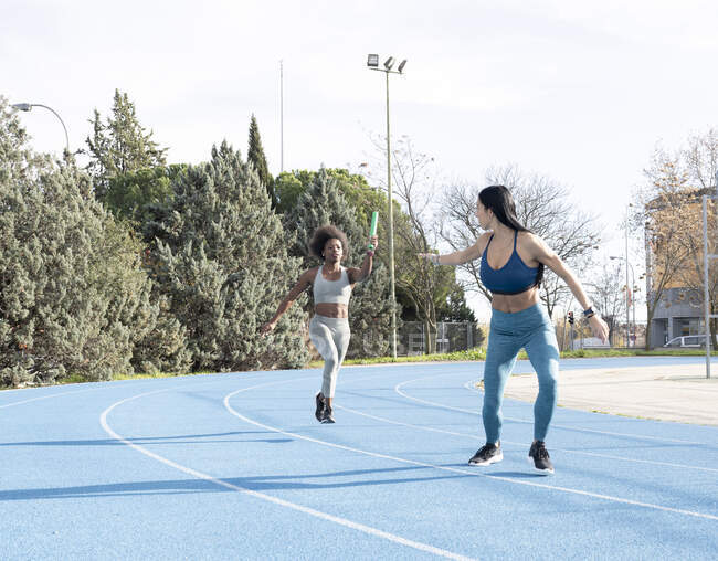 Ajuste atletas femeninos multirraciales que pasan bastón mientras corren a lo largo de la pista en el estadio durante la carrera de relevos - foto de stock