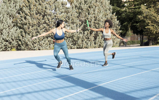 Fit multiracial female athletes passing baton while running along track at stadium during relay race — Stock Photo