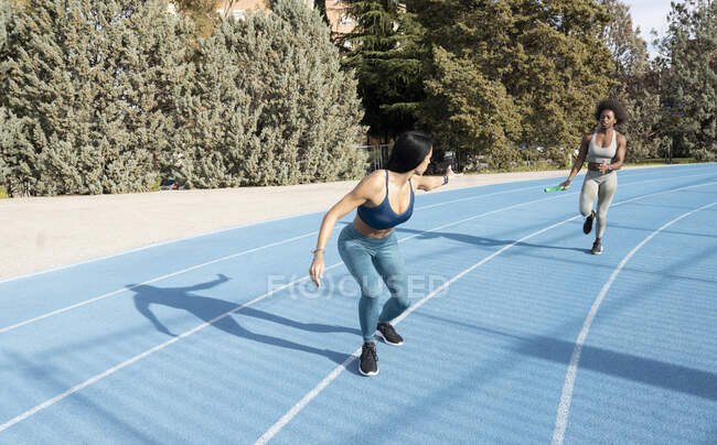 Fit multiracial female athletes passing baton while running along track at stadium during relay race — Stock Photo