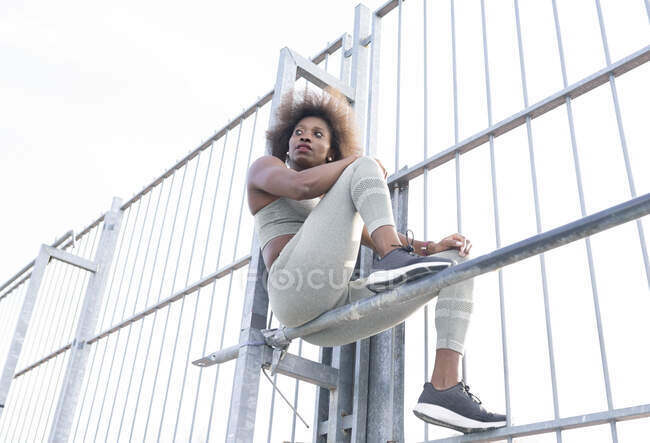 From below of African American female athlete in sportswear sitting on metal horizontal bar during training on sports ground and looking away — Stock Photo