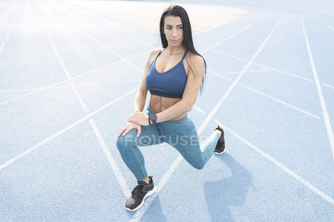 Alto ângulo de ajuste feminino corredor aquecendo no estádio e fazendo exercícios lunge enquanto se prepara para o treino no estádio e olhando para longe — Fotografia de Stock