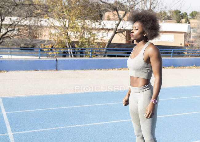 Вид сбоку концентрированного афроамериканского бегуна, стоящего на треке и готовящегося к забегу с закрытыми глазами на стадионе — стоковое фото