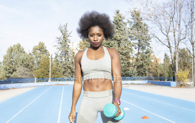 Focused black female athlete doing exercises with dumbbell while training at stadium in summer and looking forward — Stock Photo