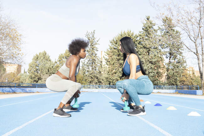 Seitenansicht diverser Sportlerinnen beim Kniebeugen mit Kurzhanteln während des Trainings im Stadion und beim gegenseitigen Betrachten — Stockfoto