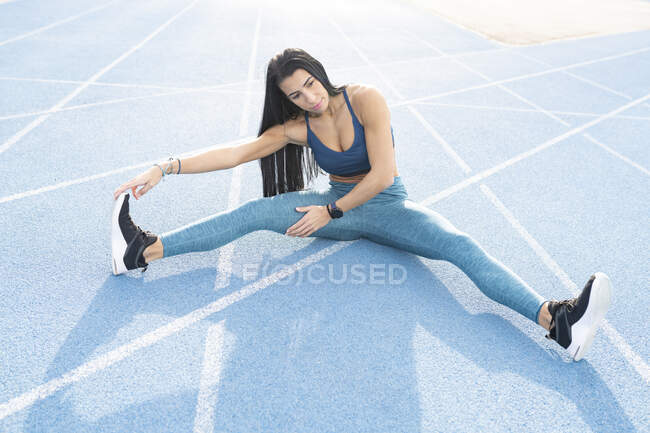 Високий кут гнучкого спортсмена, який сидить на треку і розтягує ноги, роблячи вигини і прогрівання перед тренуванням на стадіоні — стокове фото
