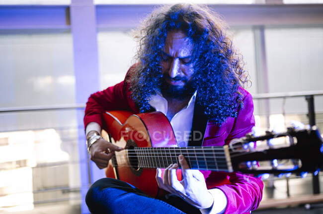 Hombre étnico enfocado con el pelo largo tocando la guitarra acústica mientras ensayan la canción en el escenario a la luz del foco - foto de stock