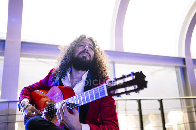 Hombre étnico enfocado con el pelo largo tocando la guitarra acústica mientras ensayan la canción en el escenario a la luz del foco - foto de stock