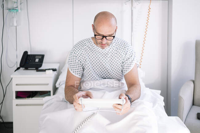 Hombre adulto concentrado que usa bata de paciente y anteojos que navegan tableta en la cama en la sala de luz en el hospital moderno - foto de stock