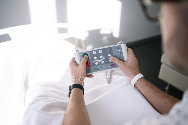 Взрослый мужчина в больничном халате и очках просматривает пульт дистанционного управления больничной койки на кровати в светлом отделении современной больницы — стоковое фото