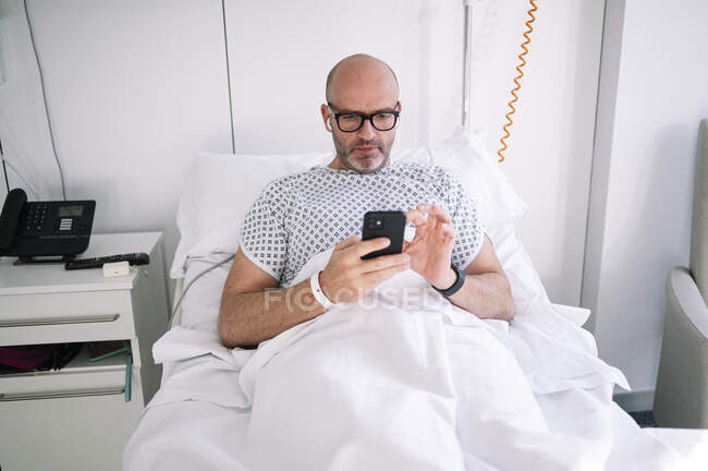 Hombre adulto concentrado que usa bata de paciente y anteojos que navegan por el teléfono en la cama en la sala de luz en el hospital moderno - foto de stock