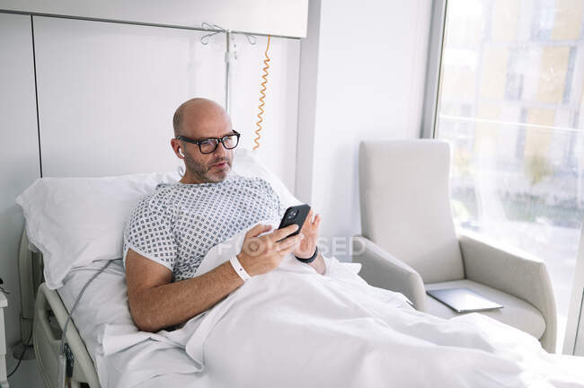 Concentrado adulto masculino vestindo vestido de paciente e óculos de navegação telefone na cama em ala de luz no hospital moderno — Fotografia de Stock