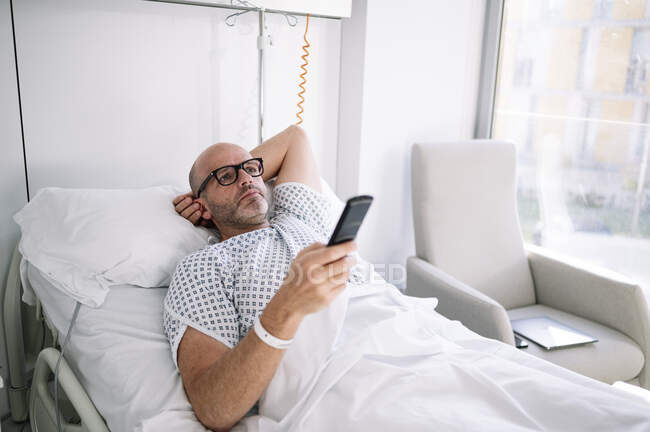 Дорослий пацієнт чоловічої статі в лікарняній сукні лежить на ліжку і використовує дистанційне керування в легко обладнаному приміщенні в клініці — стокове фото