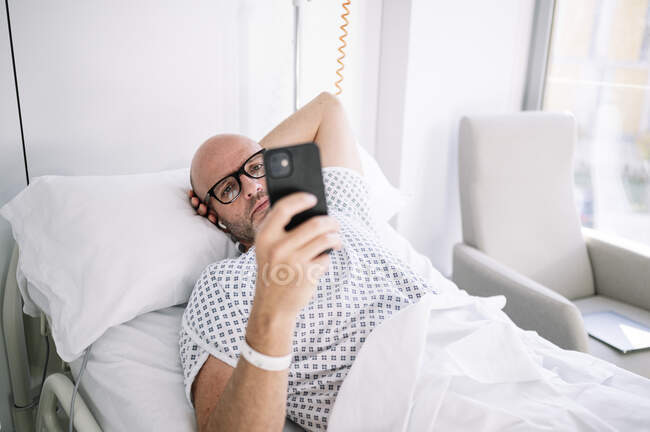 Concentrado adulto masculino vestindo vestido de paciente e óculos de navegação telefone na cama em ala de luz no hospital moderno — Fotografia de Stock