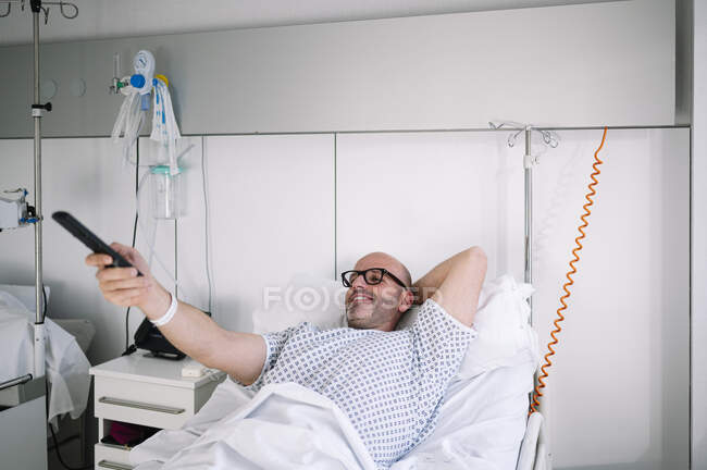 Patient adulte souriant en robe d'hôpital couché sur le lit et utilisant la télécommande dans la salle équipée de lumière dans la clinique — Photo de stock
