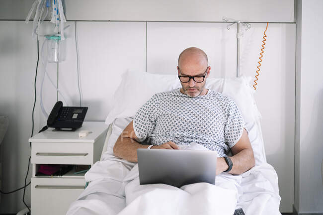 Concentré adulte patient masculin en robe d'hôpital et lunettes parcourant netbook alors qu'il était allongé sur le lit dans la salle de lumière à la clinique — Photo de stock