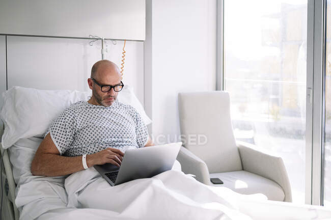 Концентрированный взрослый пациент в больничном халате и очках просматривает нетбук, лежа на кровати в светлом отделении клиники — стоковое фото