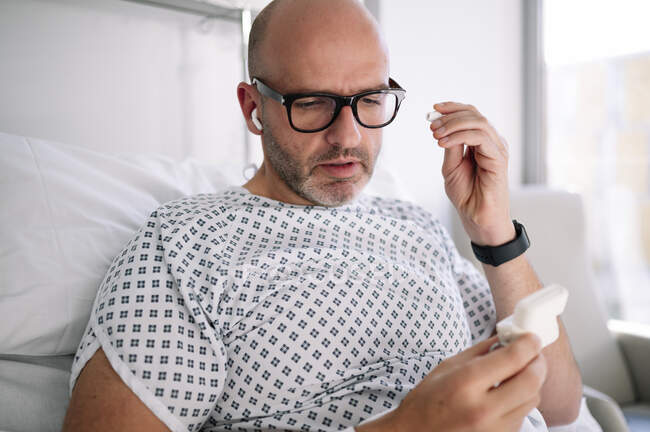 Cultivo adulto paciente masculino en bata de hospital que se pone auriculares TWS mientras está acostado en la cama en la sala moderna en la clínica - foto de stock