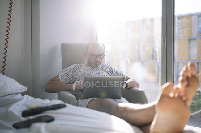 Paciente masculino adulto reflexivo en bata de hospital que navega netbook moderno mientras que se sienta en el sillón en sala equipada soleada en la clínica - foto de stock