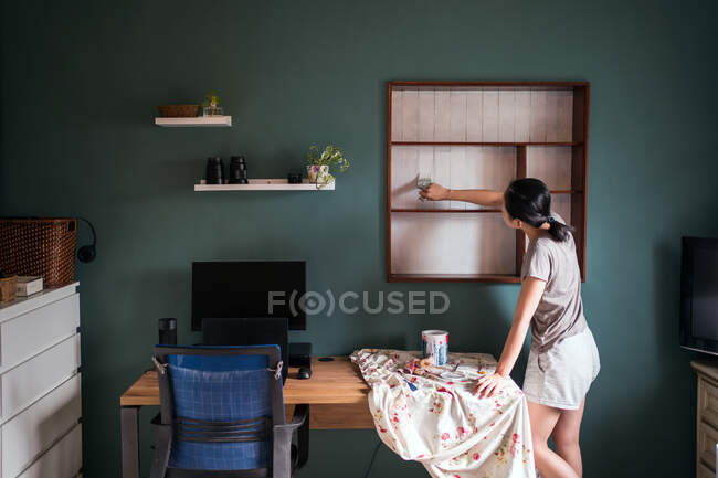 Vista laterale di femmina etnica con pennello pittura mensole in legno di colore bianco durante la ristrutturazione di mobili — Foto stock