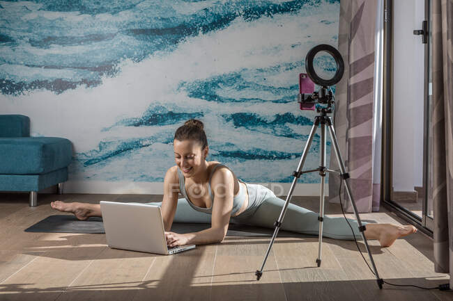 Положительная женщина фитнес-блоггер запись в прямом эфире онлайн видео на смартфоне на штативе и просмотр нетбука на полу — стоковое фото