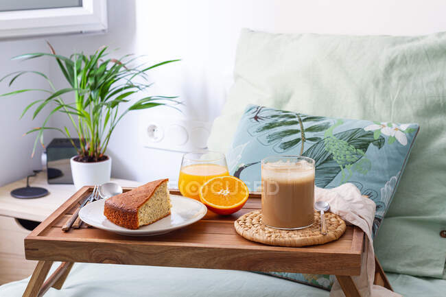 Cup of coffee and homemade sponge cake placed on wooden tray with glass of fresh orange juice prepared for breakfast in bedroom — Stock Photo