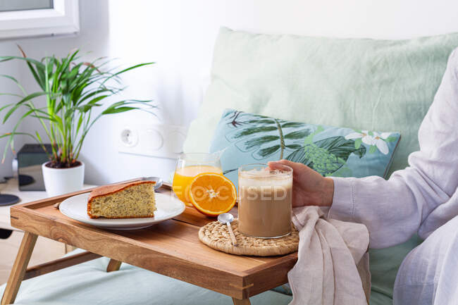 Cosecha irreconocible hembra sentada en la cama con taza de café y bandeja servida con bizcocho y vaso de jugo mientras desayunamos en casa - foto de stock