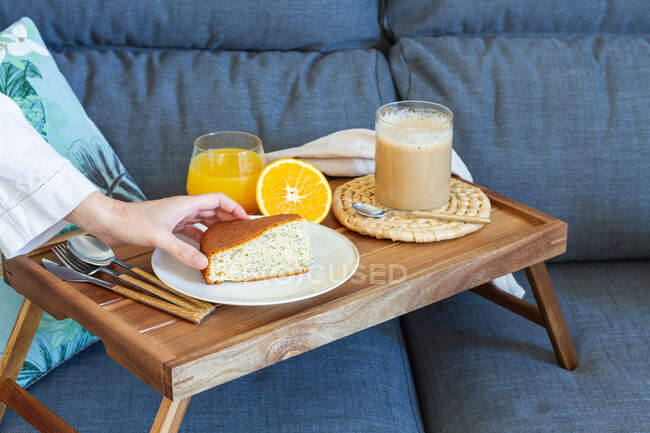 High angle of crop faceless female taking piece of homemade sponge cake from tray with cup of coffee and glass of orange juice during breakfast at home — Stock Photo